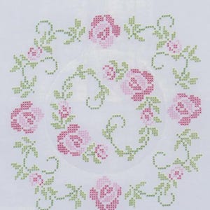 Puede incluir: Bordado de punto de cruz con un diseño floral circular. Rosas rosas y hojas verdes dispuestas en forma de corona sobre un fondo blanco. La labor muestra una estética detallada y artesanal, ideal para decoración del hogar o proyectos de manualidades.