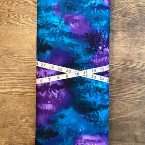 May include: A rolled fabric bolt with a vivid, abstract design in blue, purple, and black, reminiscent of a starry night or forest scene. A measuring tape shows a length of approximately 89 cm.