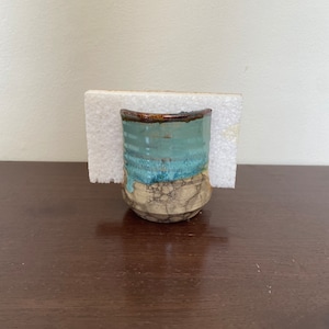 Puede incluir: Un pequeño jarrón de cerámica con un esmalte azul claro y detalles marrones. El jarrón tiene un acabado craquelado y está sentado sobre una esponja blanca.