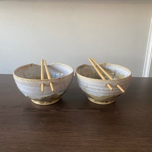 以下が含まれることがあります： 茶色と青色の釉薬がかかった白い陶器のボウルが2つ。それぞれに木製の箸が1膳ずつ入っています。