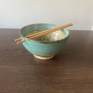 以下が含まれることがあります： 薄い青色のセラミックボウルで、茶色の縁と白い内側があります。ボウルは濃い茶色の木製の表面に置かれ、中に木製の箸が置かれています。