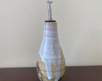 Puede incluir: Una botella de aceite de oliva de cerámica con un esmalte marrón claro y un esmalte blanco en goteo. La botella tiene un pico de vertido de acero inoxidable.