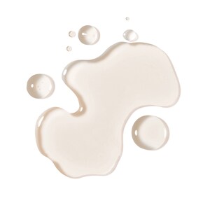 May include: A close-up shot of a light beige liquid, possibly a cosmetic product, spilled on a white background. The liquid forms an irregular shape with several smaller, round droplets scattered around it. The overall effect is clean and minimalist.