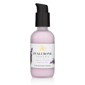 May include: A clear glass bottle with a black pump top filled with a pink liquid. The bottle has a white label with gold lettering that reads "HYALURONIC SERUM 8 Hydrates, Plumps & Targets Wrinkles LUXE HEAVENLY BODIES". There is a small image of lavender flowers on the label.