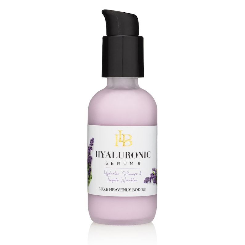 May include: A clear glass bottle with a black pump top filled with a pink liquid. The bottle has a white label with gold lettering that reads "HYALURONIC SERUM 8 Hydrates, Plumps & Targets Wrinkles LUXE HEAVENLY BODIES". There is a small image of lavender flowers on the label.