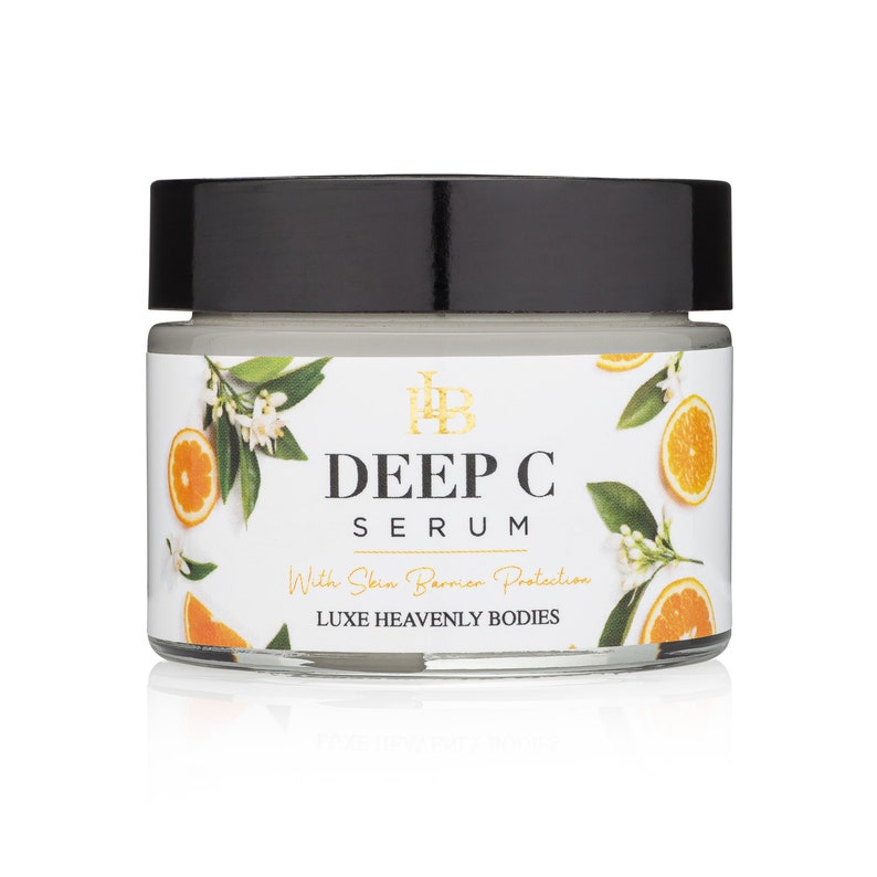 May include: A white jar with a black lid containing a white cream. The jar has a label with the text "DEEP C SERUM" and "LUXE HEAVENLY BODIES". The label also includes the text "With Skin Barrier Protection". The jar is surrounded by white flowers and orange slices.