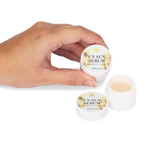 May include: White sample-size jars of "UN SUN SERUM" with sunflower and text design. One jar is open, revealing a light-colored serum. The jars are held by a hand against a white background. The website address is also visible.