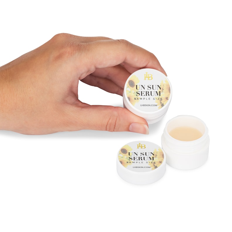 May include: White sample-size jars of "UN SUN SERUM" with sunflower and text design. One jar is open, revealing a light-colored serum. The jars are held by a hand against a white background. The website address is also visible.