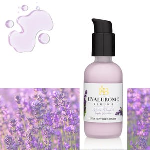 May include: A clear glass bottle with a black pump top filled with a pink liquid. The bottle is labeled "HYALURONIC SERUM" and "LUXE HEAVENLY BODIES". The bottle is sitting on a white surface with a lavender flower background.
