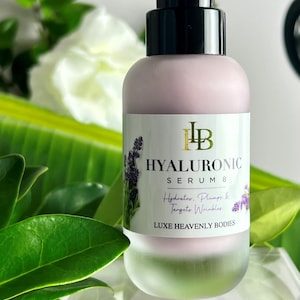 May include: A clear glass bottle with a black pump top and a frosted pink label. The label reads "HYALURONIC SERUM 8" and "Hydrator, Plumper & Targets Wrinkles". The bottle is sitting on a clear glass surface with green leaves in the background. The label also includes the text "LUXE HEAVENLY BODIES".