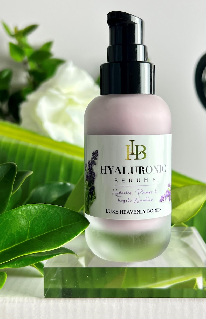 May include: A clear glass bottle with a black pump top and a frosted pink label. The label reads "HYALURONIC SERUM 8" and "Hydrator, Plumper & Targets Wrinkles". The bottle is sitting on a clear glass surface with green leaves in the background. The label also includes the text "LUXE HEAVENLY BODIES".