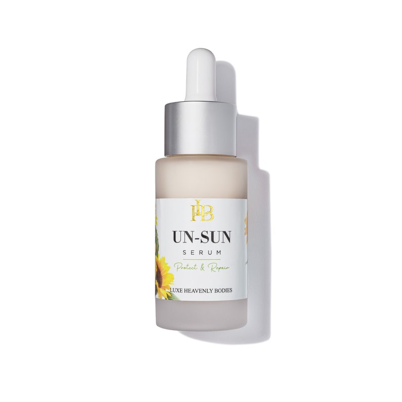 May include: A frosted glass bottle of UN-SUN Serum with a silver cap and dropper. The label features the text "UN-SUN SERUM Protect & Repair" with a sunflower illustration. The product is from LUXE HEAVENLY BODIES.