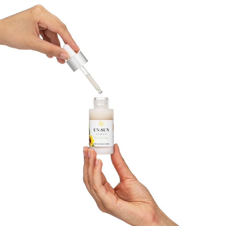 May include: A white serum bottle with a dropper, held by two hands against a white background. The bottle has a label with the text "UN-SUN SERUM" and a sunflower graphic. The dropper is silver and clear, with a drop of serum falling.