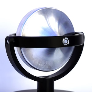 May include: A silver-colored metal sphere mounted within a black circular frame that is supported by a black cylindrical post and a flat, circular black base. The frame features a small, circular silver accent on the side of the ring.