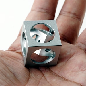 Puede incluir: Un cubo de metal de color plateado con un gran recorte circular en cada una de sus seis caras, que revela un cubo sólido más pequeño suspendido en el centro de la estructura. El objeto tiene un acabado mate liso y un diseño geométrico.