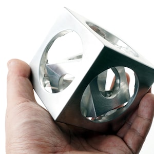 May include: A silver-colored metal cube sculpture featuring large circular cutouts on each face, revealing a smaller, nested geometric shape inside. The object has a smooth, brushed metallic finish and a hollow, open-frame construction.