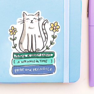 Puede incluir: Un gato blanco con los ojos cerrados se sienta encima de una pila de tres libros. Los libros se titulan "Anne of Green Gables", "A Wrinkle in Time" y "Pride and Prejudice". El gato está rodeado de flores amarillas.