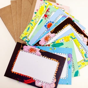 May include: A stack of colorful blank note cards with floral borders. The cards are arranged in a fan shape and have a white background with a black border.