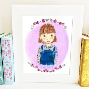 May include: A framed illustration of a young girl with brown hair wearing a blue overall dress and a plaid shirt. The illustration is surrounded by a wreath of pink flowers and the text "Scout Finch" is written below the girl.