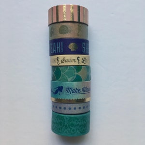 May include: A stack of eight washi tapes with various designs and colors, including gold, pink, blue, green, and white. The tapes feature mermaid silhouettes, seashells, and the text "Make Waves" and "Swim & Shine".
