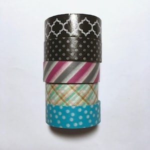 May include: A stack of five rolls of washi tape. The top roll has a black and white geometric pattern. The second roll has a black and white polka dot pattern. The third roll has a pink, gray, and white striped pattern. The fourth roll has a green, gold, and white plaid pattern. The bottom roll has a blue and white polka dot pattern.