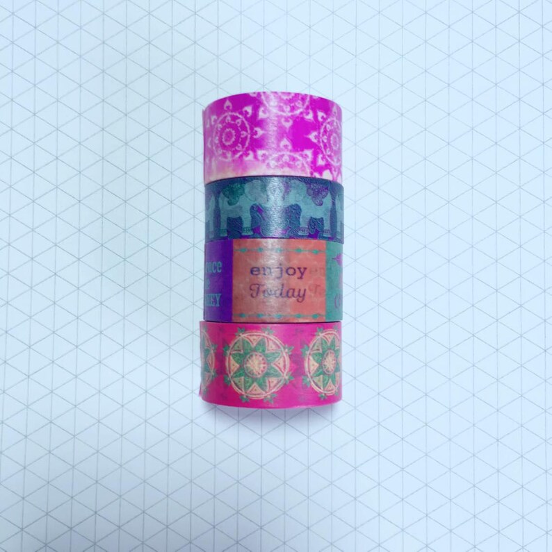May include: Four rolls of washi tape stacked on top of each other. The top roll is pink with a white mandala design. The second roll is purple with a cityscape design. The third roll is orange with the text "enjoy today". The bottom roll is pink with a green mandala design.