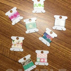 May include: Eight spools of colorful washi tape on white plastic spools. The washi tape has various patterns including polka dots, geometric shapes, and floral designs.