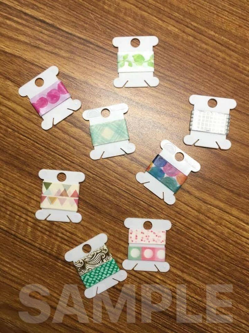 May include: Eight spools of colorful washi tape on white plastic spools. The washi tape has various patterns including polka dots, geometric shapes, and floral designs.