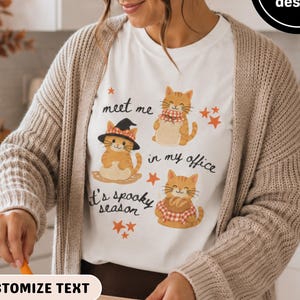 May include: A white t-shirt featuring three orange tabby cat illustrations, orange stars, and the text 'meet me in my office' and 'it's spooky season'. One cat wears a black witch hat, one wears a scarf, and one holds a loaf of bread.