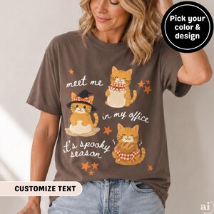 May include: A dark brown t-shirt featuring three orange tabby cats and orange star graphics. The text reads "meet me in my office" and "it's spooky season". One cat wears a black witch hat, another wears a red and white checkered scarf.