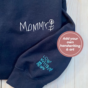 May include: Navy blue sweatshirt with white embroidered text reading "Mommy" and a stick figure. The sleeve features teal embroidery with the words "Love Matthew and Jackson". A circular graphic says "Add your own handwriting & art".