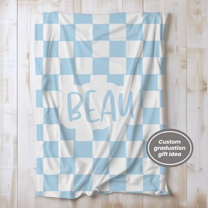 May include: A soft, light blue and white checkered blanket with the name "BEAU" in light blue script. The blanket is draped over a light wood surface. A circular graphic in the bottom right corner reads "Custom graduation gift idea."