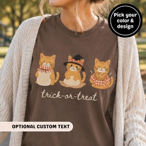 May include: A brown t-shirt featuring a graphic of three orange tabby cats with the text "trick-or-treat" below them. One cat wears a witch hat, while the others hold baked goods. The shirt is layered under a cream-colored knit cardigan.