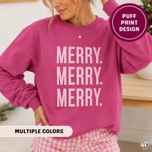 May include: A bright magenta long-sleeve crewneck sweatshirt featuring the text MERRY. MERRY. MERRY. in a light pink puff print design on the front. The garment has a ribbed neckline, cuffs, and hem, and is paired with pink and white gingham shorts.