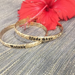 Pu&ograve; includere: Due braccialetti dorati incisi con motivi floreali e i nomi "Kalene" e "Jade Skye" in lettere nere. Un vivace fiore di ibisco rosso &egrave; sullo sfondo, su una superficie grigia strutturata.
