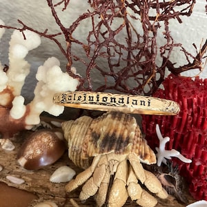Pu&ograve; includere: Un braccialetto rigido color oro con l'iscrizione "Kalejuionalani" in un carattere decorativo. Il braccialetto &egrave; esposto tra conchiglie, coralli e alghe secche, creando un'estetica da spiaggia.