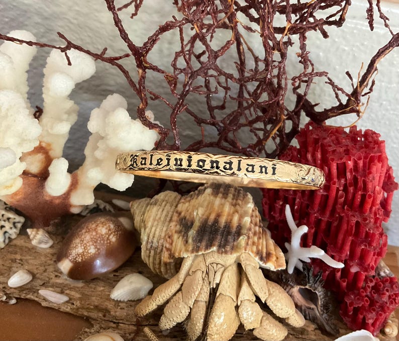 Pu&ograve; includere: Un braccialetto rigido color oro con l'iscrizione "Kalejuionalani" in un carattere decorativo. Il braccialetto &egrave; esposto tra conchiglie, coralli e alghe secche, creando un'estetica da spiaggia.