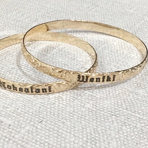 Pu&ograve; includere: Due braccialetti color oro con motivi floreali incisi e testo. Un braccialetto recita "Nohealani" e l'altro "Weniki" in un carattere scuro. I braccialetti sono esposti su una superficie strutturata bianco sporco.