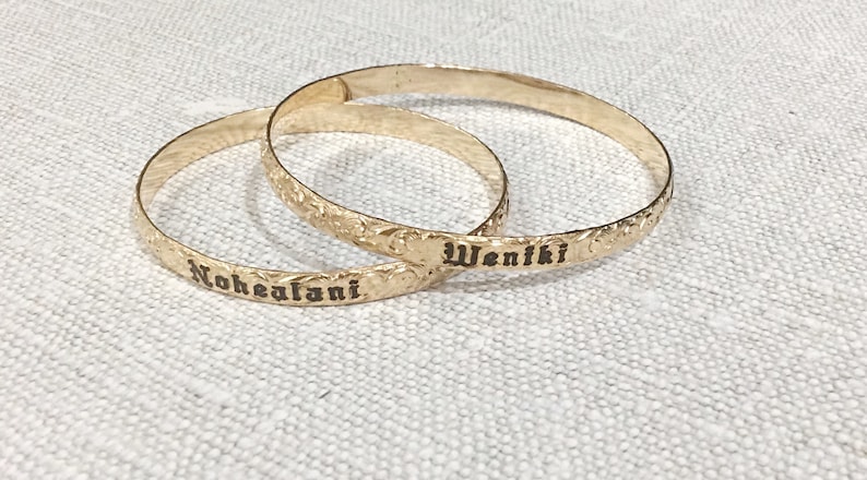Pu&ograve; includere: Due braccialetti color oro con motivi floreali incisi e testo. Un braccialetto recita "Nohealani" e l'altro "Weniki" in un carattere scuro. I braccialetti sono esposti su una superficie strutturata bianco sporco.