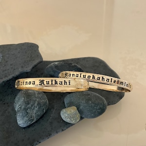 Pu&ograve; includere: Due braccialetti color oro con testo inciso e motivi floreali. Un braccialetto recita "Rainoa Kuikahi" e l'altro "Kanaluokahaleimiala". I gioielli sono esposti su pietre grigio scuro.