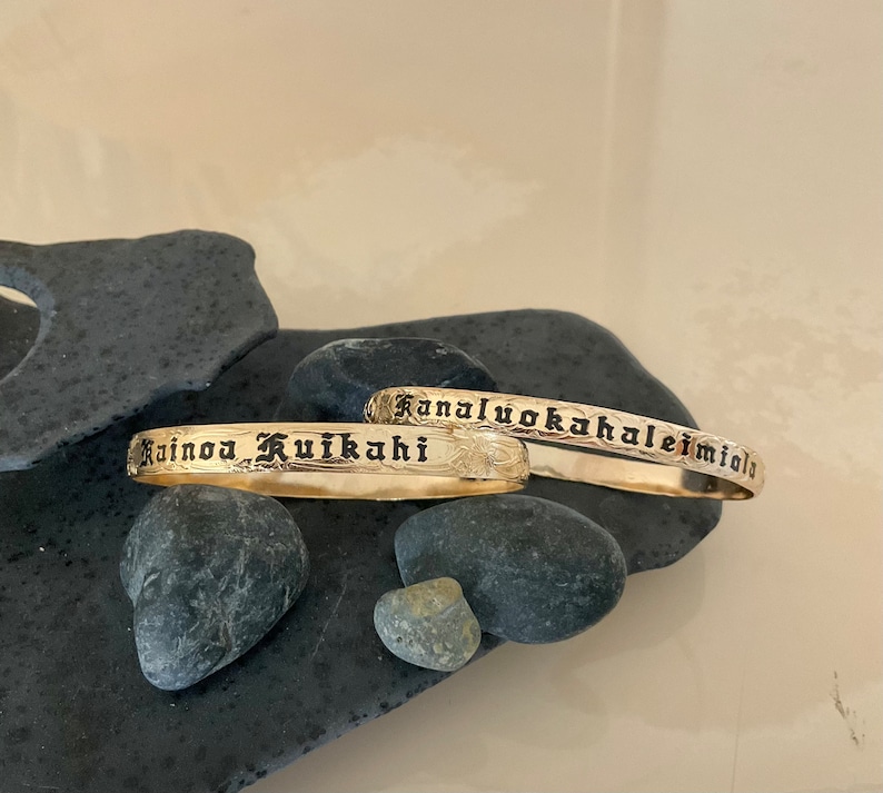 Pu&ograve; includere: Due braccialetti color oro con testo inciso e motivi floreali. Un braccialetto recita "Rainoa Kuikahi" e l'altro "Kanaluokahaleimiala". I gioielli sono esposti su pietre grigio scuro.