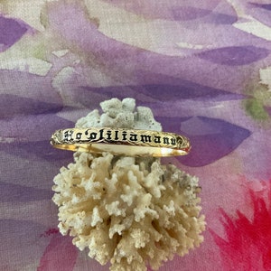 Pu&ograve; includere: Un braccialetto color oro con l'iscrizione "Ho'oliliamann" in lettere nere. Il braccialetto &egrave; appoggiato su corallo bianco su uno sfondo colorato di viola, rosa e rosso.