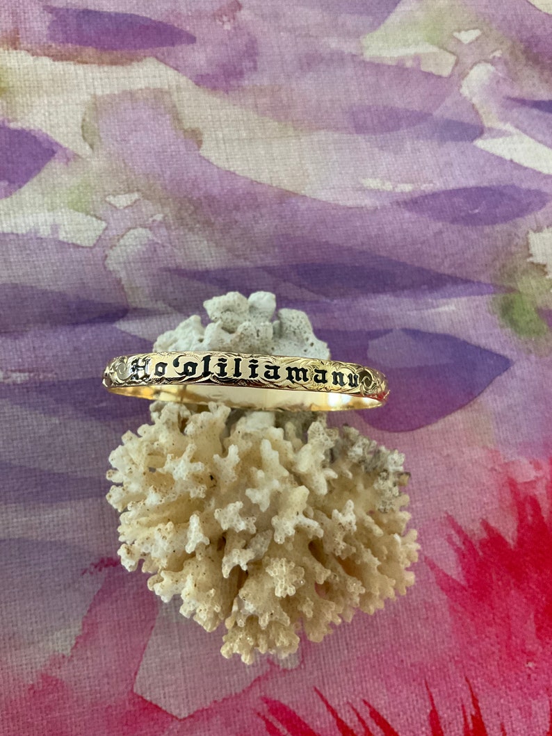 Pu&ograve; includere: Un braccialetto color oro con l'iscrizione "Ho'oliliamann" in lettere nere. Il braccialetto &egrave; appoggiato su corallo bianco su uno sfondo colorato di viola, rosa e rosso.