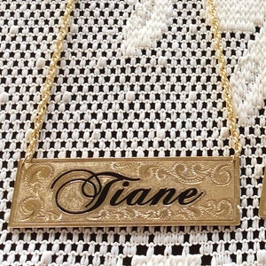 May include: Gold-colored metal bottle tag with a chain. The tag has an engraved design and the name "Tiane" in black lettering.