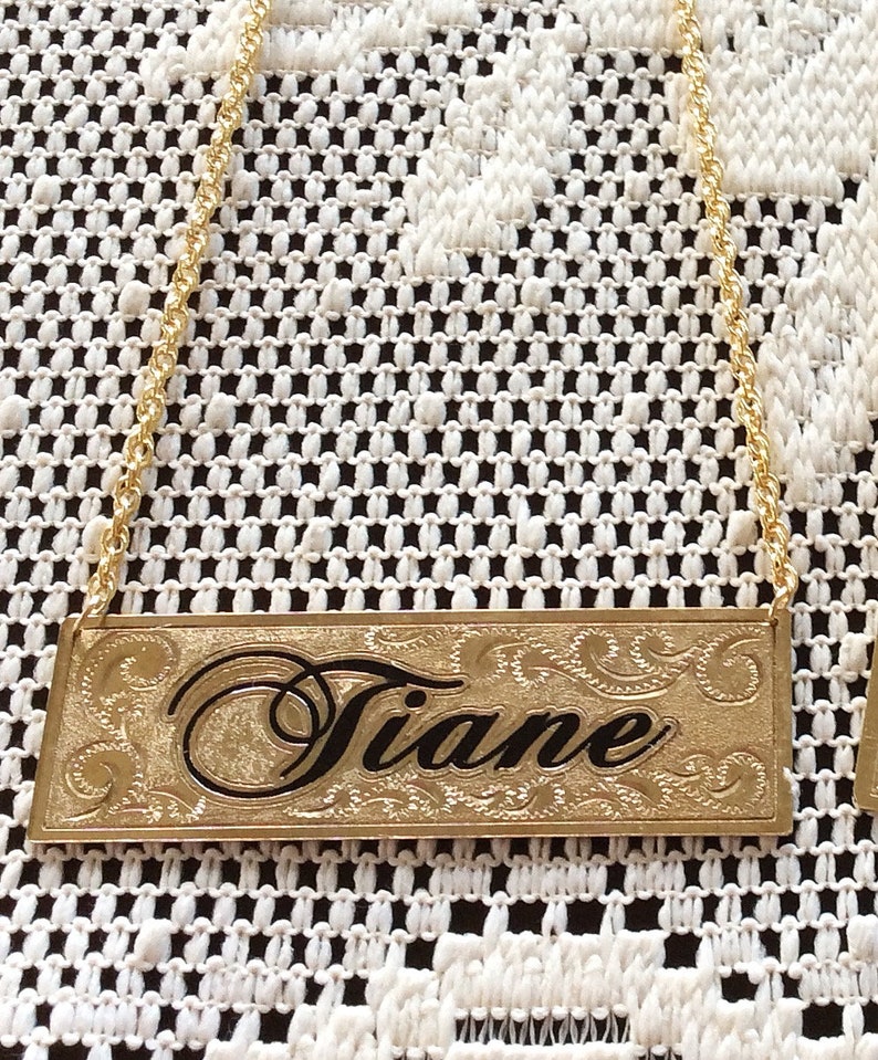 May include: Gold-colored metal bottle tag with a chain. The tag has an engraved design and the name "Tiane" in black lettering.