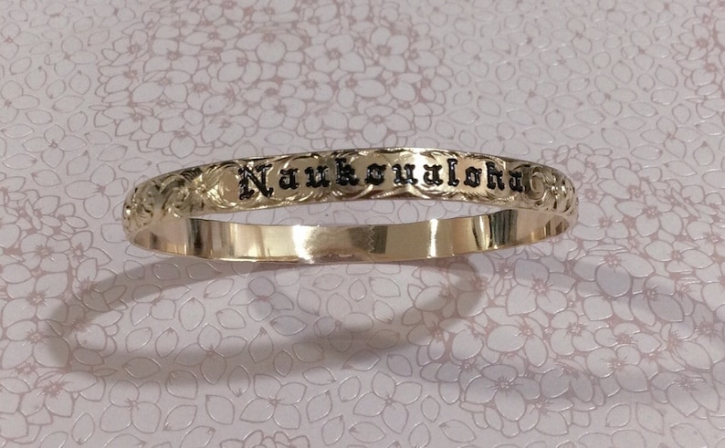 Pu&ograve; includere: Un braccialetto a cerchio color oro con la parola "Naukoaloha" incisa in lettere nere. Il braccialetto presenta un motivo floreale attorno al testo. Lo sfondo &egrave; un motivo floreale rosa chiaro.