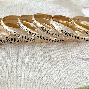 Pu&ograve; includere: Braccialetti dorati con nomi incisi in lettere nere. I braccialetti presentano un motivo floreale sui bordi. I nomi includono "Jez", "Olivia June", "Latticia", "Kauai Inoahaneheokalani", "Leilani" e "Kalamakūokalani".