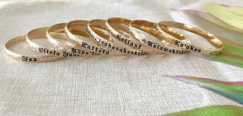 Pu&ograve; includere: Braccialetti dorati con nomi incisi in lettere nere. I braccialetti presentano un motivo floreale sui bordi. I nomi includono "Jez", "Olivia June", "Latticia", "Kauai Inoahaneheokalani", "Leilani" e "Kalamakūokalani".