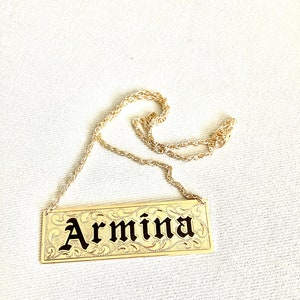May include: Gold necklace with a rectangular nameplate pendant that says "Armina" in black lettering.