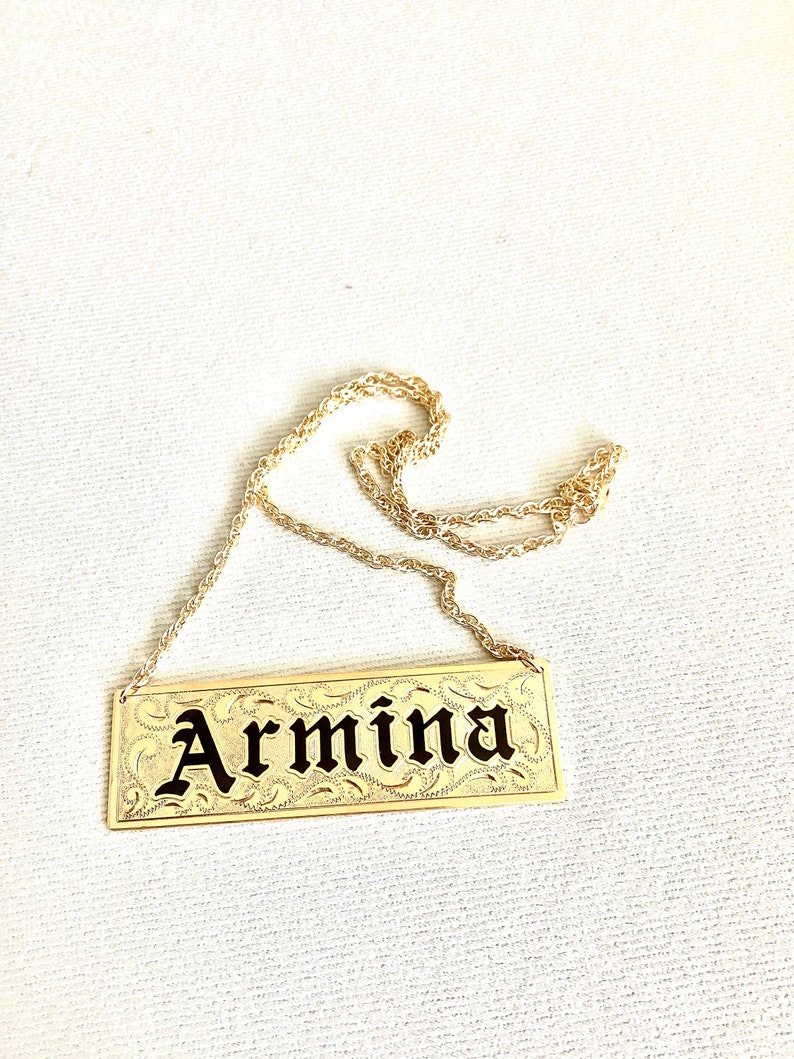 May include: Gold necklace with a rectangular nameplate pendant that says "Armina" in black lettering.